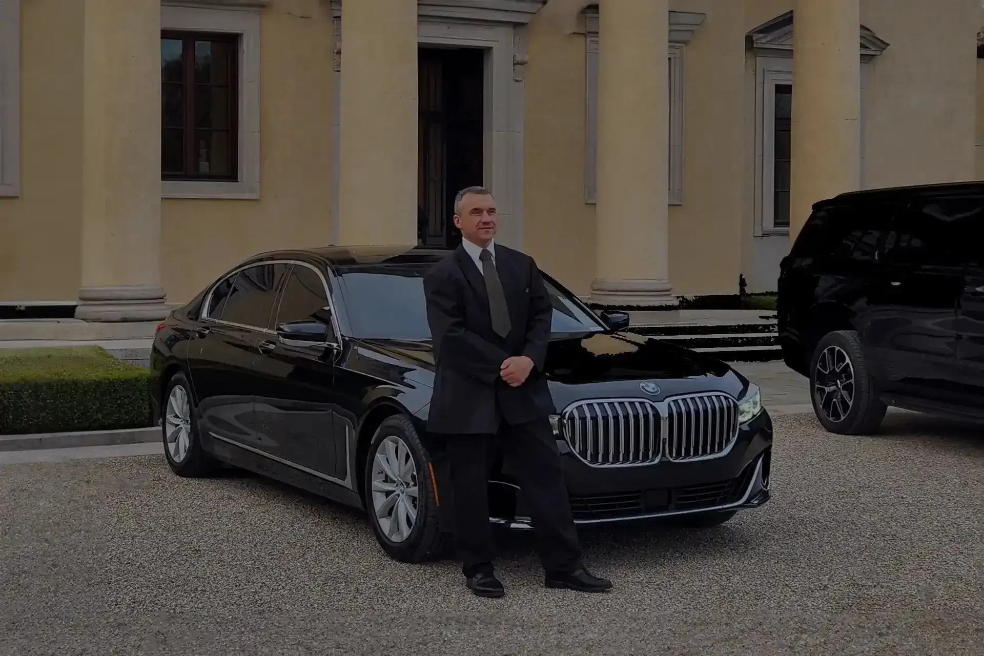 A formally attired chauffeur stands proudly beside a sleek, black Mercedes-Benz sedan, showcasing Affects Premium